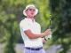 Golfer in a white polo and hat watches the ball mid-swing on a sunny course with a golf club raised high.