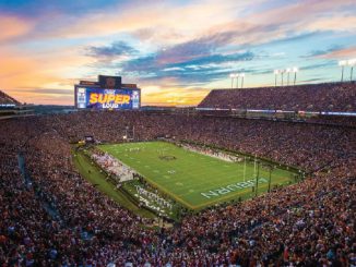 Packed football stadium at sunset, players on the bright green field, and a huge crowd with a screen reading 'SUPER LOUD.'
