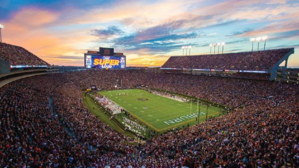 Packed football stadium at sunset, players on the bright green field, and a huge crowd with a screen reading 'SUPER LOUD.'