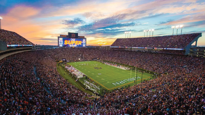 Packed football stadium at sunset, players on the bright green field, and a huge crowd with a screen reading 'SUPER LOUD.'