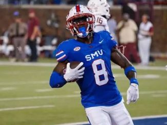 SMU football player wearing blue jersey number 8 runs with the ball during a game, white pants, on a green field with spectators in the background.