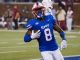 SMU football player wearing blue jersey number 8 runs with the ball during a game, white pants, on a green field with spectators in the background.