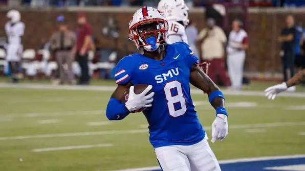 SMU football player wearing blue jersey number 8 runs with the ball during a game, white pants, on a green field with spectators in the background.