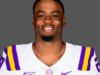 Portrait of an LSU football player in a white jersey with purple and gold stripes, smiling at the camera against a gray background.