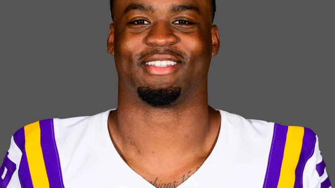 Portrait of an LSU football player in a white jersey with purple and gold stripes, smiling at the camera against a gray background.