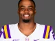 Portrait of an LSU football player in a white jersey with purple and gold stripes, smiling at the camera against a gray background.