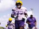 LSU football players in purple uniforms sprint across a sunny practice field, leading with player wearing number 77 and a yellow helmet