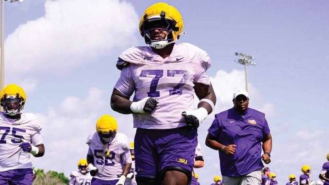 LSU football players in purple uniforms sprint across a sunny practice field, leading with player wearing number 77 and a yellow helmet
