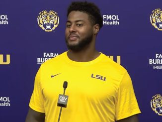Athlete in a yellow LSU shirt speaks at a press conference with a purple backdrop and microphone in front of him.