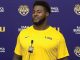 Athlete in a yellow LSU shirt speaks at a press conference with a purple backdrop and microphone in front of him.