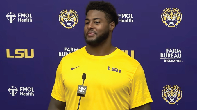 Athlete in a yellow LSU shirt speaks at a press conference with a purple backdrop and microphone in front of him.