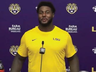 Athlete in a yellow LSU shirt speaking at a press conference with LSU and sponsor logos in the background.