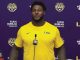 Athlete in a yellow LSU shirt speaking at a press conference with LSU and sponsor logos in the background.