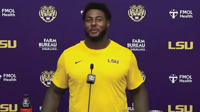 Athlete in a yellow LSU shirt speaking at a press conference with LSU and sponsor logos in the background.