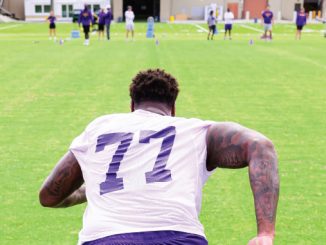 Football player seen from behind wearing a white jersey with number 77 on a practice field, teammates in the distance.