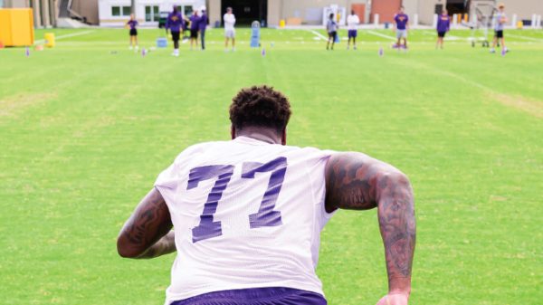 Football player seen from behind wearing a white jersey with number 77 on a practice field, teammates in the distance.