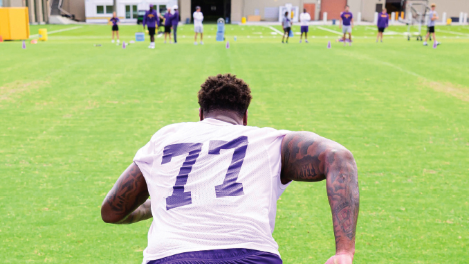 Football player seen from behind wearing a white jersey with number 77 on a practice field, teammates in the distance.