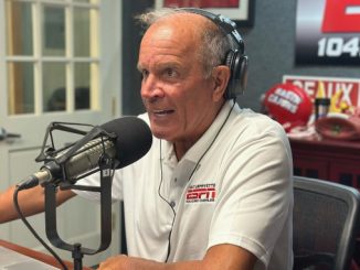 Older man wearing a headset sits at a radio studio desk, talking into a large microphone with sports memorabilia in the background.