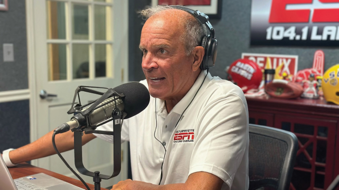 Older man wearing a headset sits at a radio studio desk, talking into a large microphone with sports memorabilia in the background.