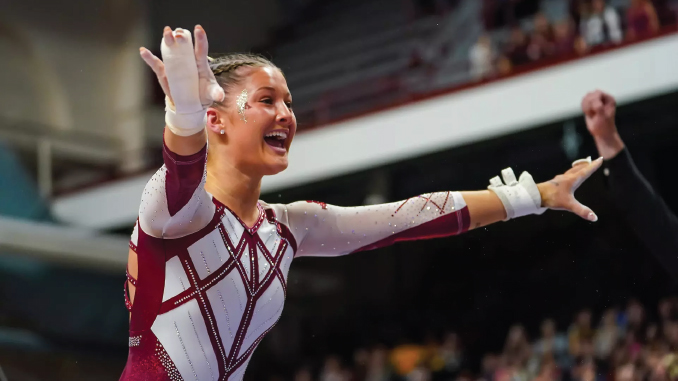 Female gymnast in a maroon and white geometric leotard raises arms in celebration during a competition, smiling at the crowd.