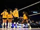 Volleyball players in yellow uniforms huddle on the court near the net during a match.