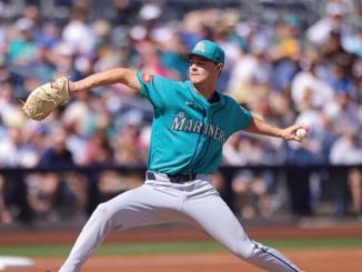 Baseball pitcher in Mariners teal uniform delivering a pitch on the mound with a blurred crowd in the stands.