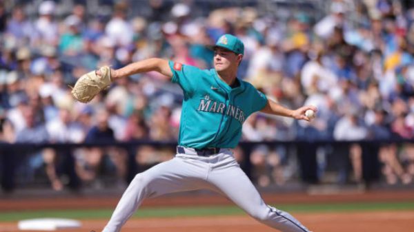 Baseball pitcher in Mariners teal uniform delivering a pitch on the mound with a blurred crowd in the stands.