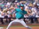 Baseball pitcher in Mariners teal uniform delivering a pitch on the mound with a blurred crowd in the stands.