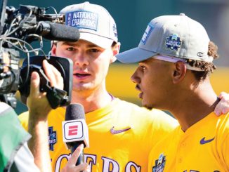 Two baseball players in yellow uniforms are interviewed by a reporter holding an ESPN microphone; both wear National Champions hats.
