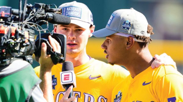 Two baseball players in yellow uniforms are interviewed by a reporter holding an ESPN microphone; both wear National Champions hats.