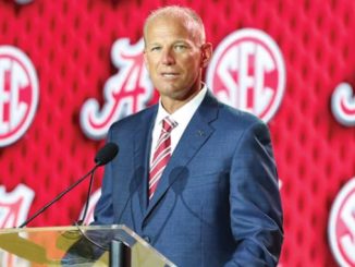 Man in a blue suit giving a speech at a podium with an SEC-branded red backdrop, speaking into a mic.