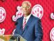Man in a blue suit giving a speech at a podium with an SEC-branded red backdrop, speaking into a mic.