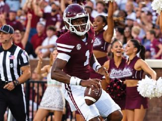 College football player wearing maroon jersey #1 runs with the ball as cheerleaders and fans celebrate in the background.
