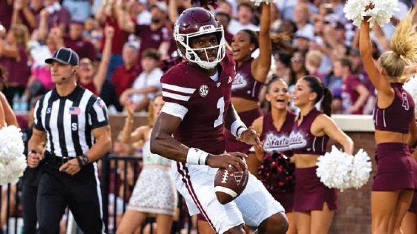 College football player wearing maroon jersey #1 runs with the ball as cheerleaders and fans celebrate in the background.