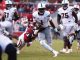 Running back in white with the ball evades red-clad defenders during a college football play.
