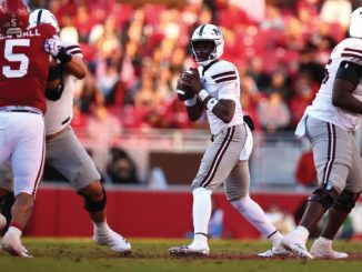 Quarterback in a white uniform drops back to throw as teammates block and red-clad players approach on the field.
