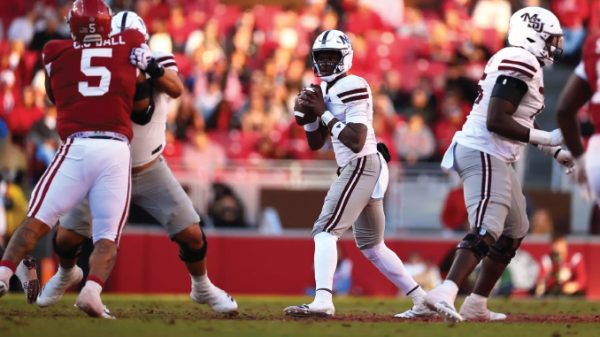 Quarterback in a white uniform drops back to throw as teammates block and red-clad players approach on the field.