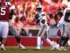 Quarterback in a white uniform drops back to throw as teammates block and red-clad players approach on the field.
