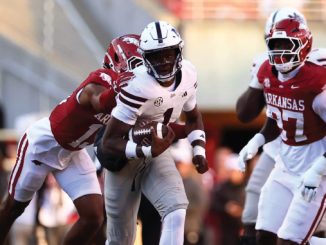 Running back in white armor carries the football while two Arkansas defenders in red chase him down the field on a sunny game day.