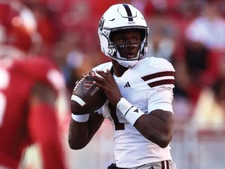 Quarterback in a white uniform holding a football, poised to throw, on a football field with red-clad defenders nearby.