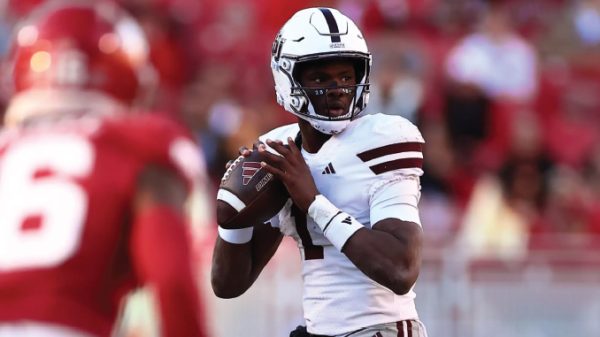 Quarterback in a white uniform holding a football, poised to throw, on a football field with red-clad defenders nearby.