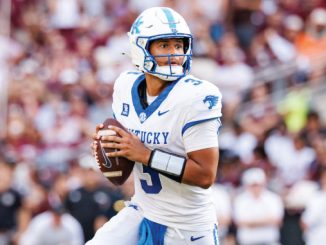 Kentucky quarterback in white and blue jersey #3 holding the football and looking downfield during a game.