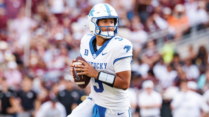 Kentucky quarterback in white and blue jersey #3 holding the football and looking downfield during a game.
