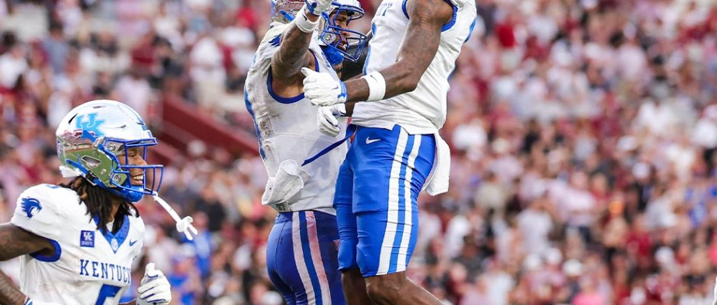 Kentucky football players in white and blue celebrate mid-air on the field as fans cheer in the packed stadium behind them.