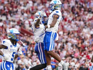 Kentucky football players in white and blue celebrate mid-air on the field as fans cheer in the packed stadium behind them.