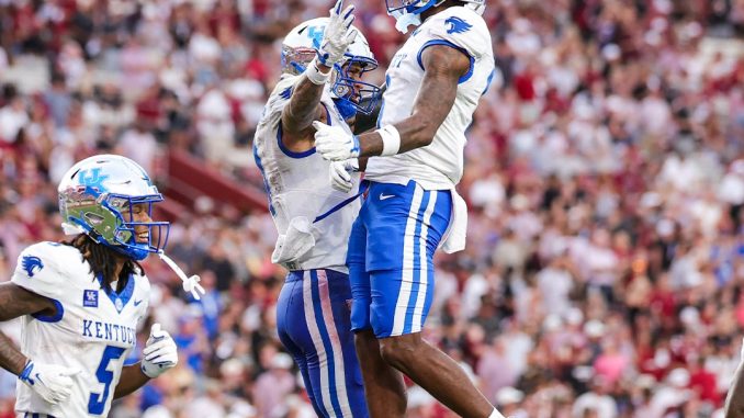 Kentucky football players in white and blue celebrate mid-air on the field as fans cheer in the packed stadium behind them.