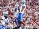 Kentucky football players in white and blue celebrate mid-air on the field as fans cheer in the packed stadium behind them.