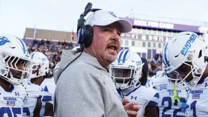 Football coach wearing a headset and gray hoodie speaks to Georgia State Panthers players on the sideline.