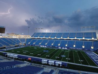 Empty Kentucky Wildcats football stadium with blue seats, a green field, and a stormy sky with lightning overhead.