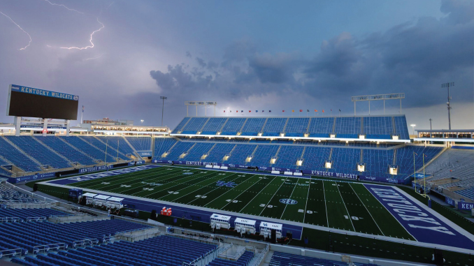 Empty Kentucky Wildcats football stadium with blue seats, a green field, and a stormy sky with lightning overhead.